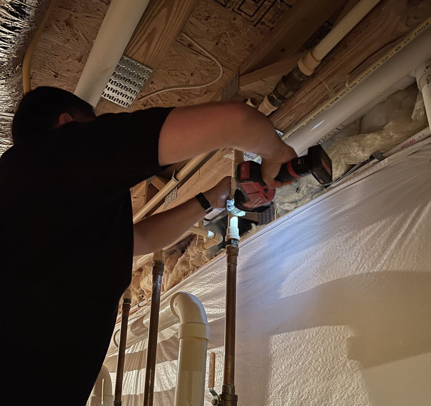 Plumber repairing water line under a house, tightening PVC and copper pipes with a cordless tool near exposed insulation