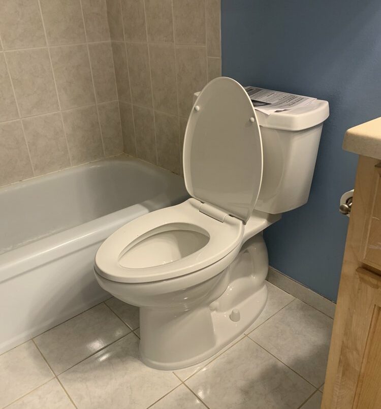 White toilet with lid and seat raised next to bathtub in tiled bathroom, paperwork on tank