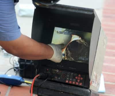 Technician viewing sewer pipe inspection video on portable monitor with gloved hand adjusting controls