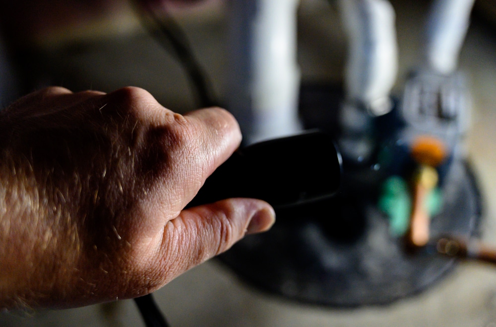 Hand holding a flashlight shining into a basement sump pump pit with PVC pipes and pump components in low light