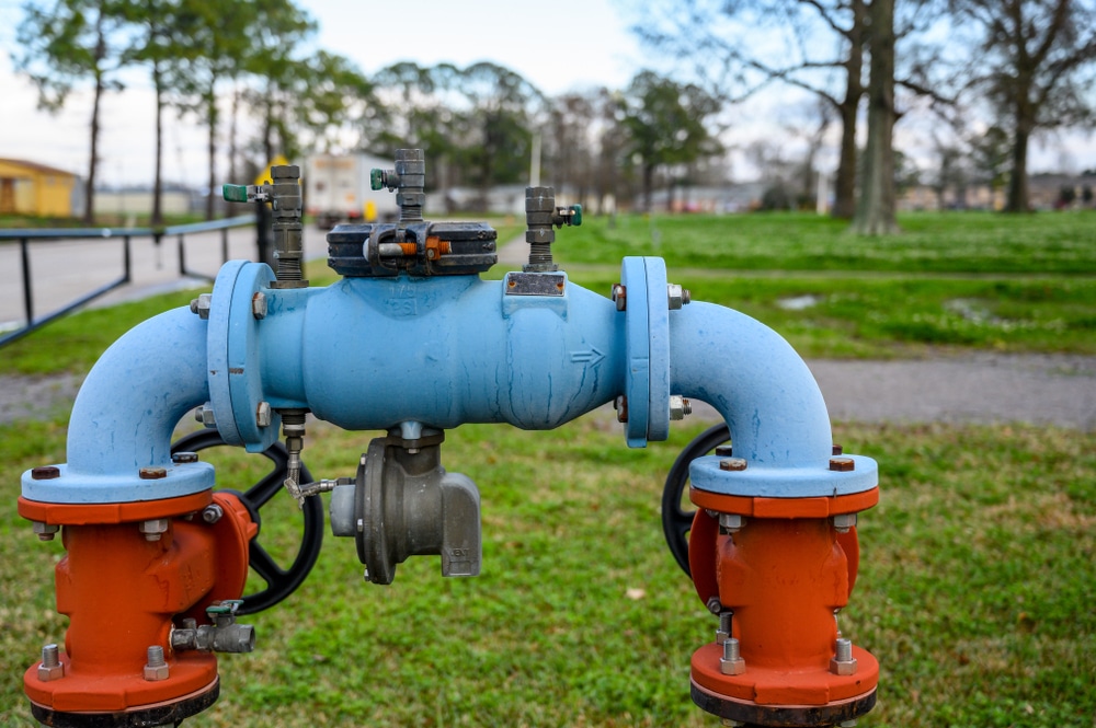 Blue pipe with meter, double block valves, and bleed valve mounted between orange flanged posts in grassy outdoor area