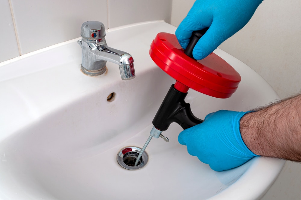 Gloved hands using a red drain auger to unclog a bathroom sink drain next to a chrome faucet