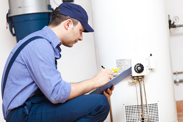 Plumber in blue uniform inspecting a water heater and taking notes on a clipboard in a utility room