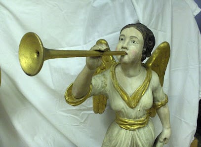 Painted angel sculpture with gold wings blowing a long brass trumpet, photographed against a white cloth backdrop