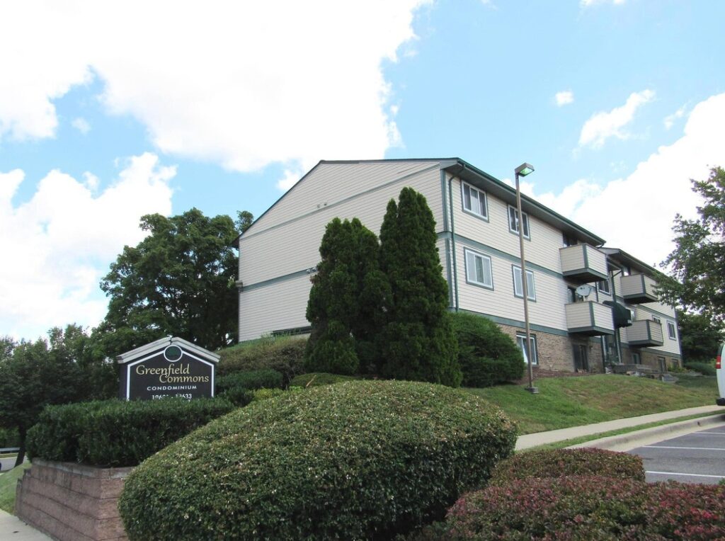 Greenfield Commons condominium building with balconies on a grassy hill, entrance sign, shrubs, and parking lot under blue sky