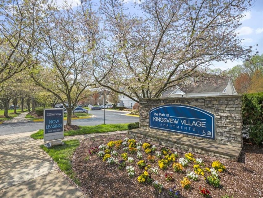 Kingsview Village apartments entrance sign by stone wall, spring trees and flower bed, with “Now Leasing” sign near sidewalk