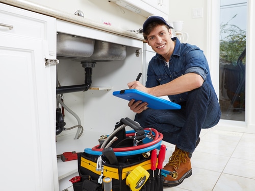 Local plumber inspecting and writing notes under a kitchen sink with tool bag and hoses nearby