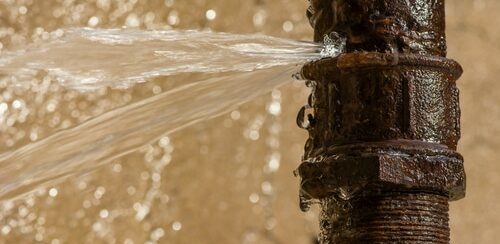 Water spraying from cracked, rusted metal pipe fitting, indicating a leaking water line needing repair