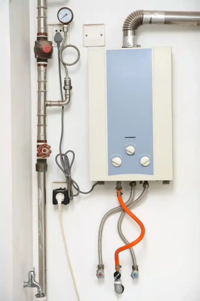 Wall-mounted tankless water heater with exposed pipes, valves, pressure gauge, and flexible water lines connected below