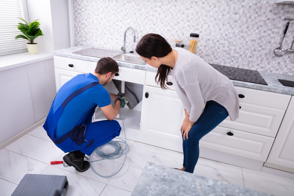 Plumber repairing garbage disposal under kitchen sink while homeowner watches, tools and drain cable on floor