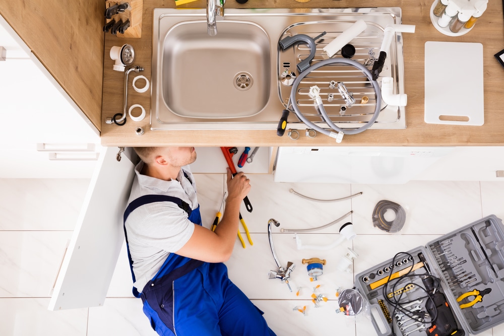 High-angle view of plumber in blue overalls repairing kitchen sink with tools and parts spread on floor