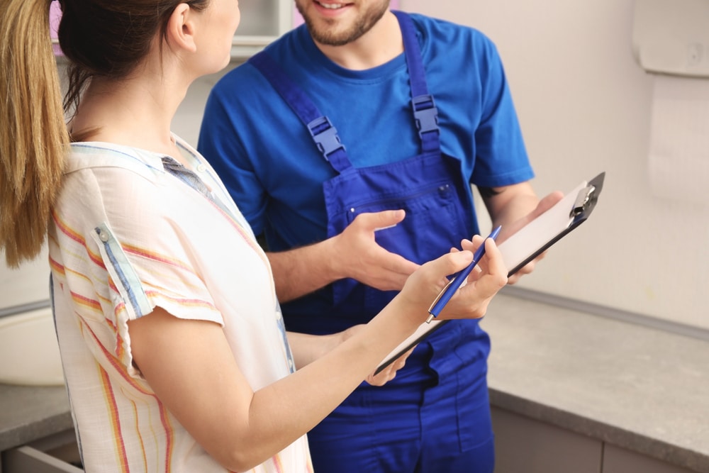 Plumber in blue uniform talks with client indoors as she holds a clipboard and pen, discussing service details