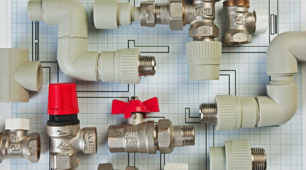 Assorted plumbing fixtures including PVC elbows, threaded pipe fittings, and metal ball valves on a blueprint-style grid background
