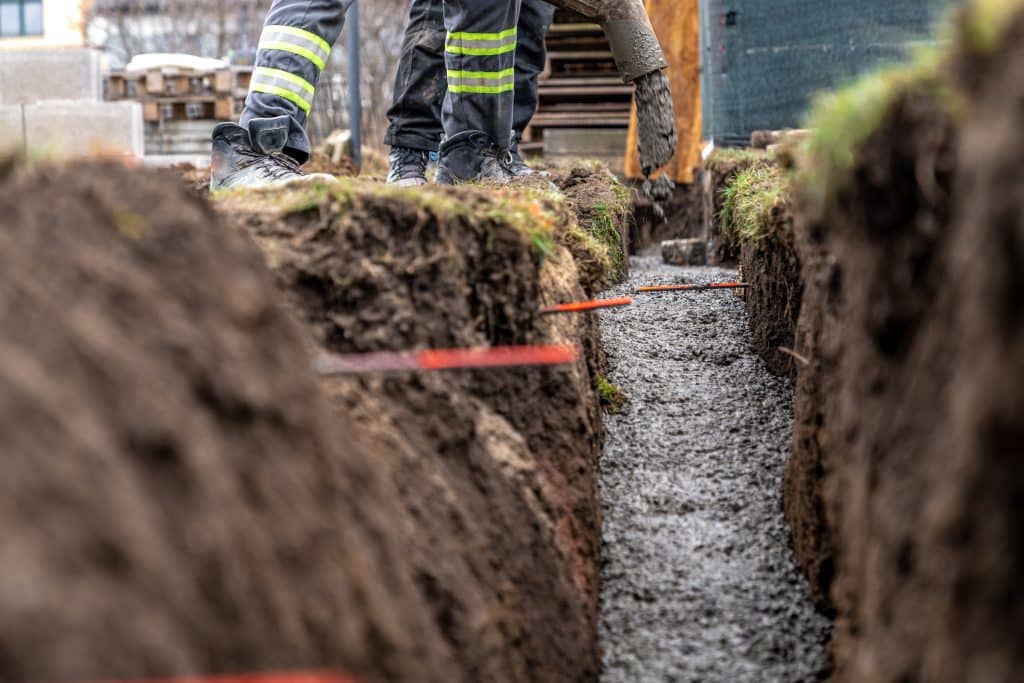 Concrete being poured into a narrow trench for a building foundation, with workers’ boots visible along the edge