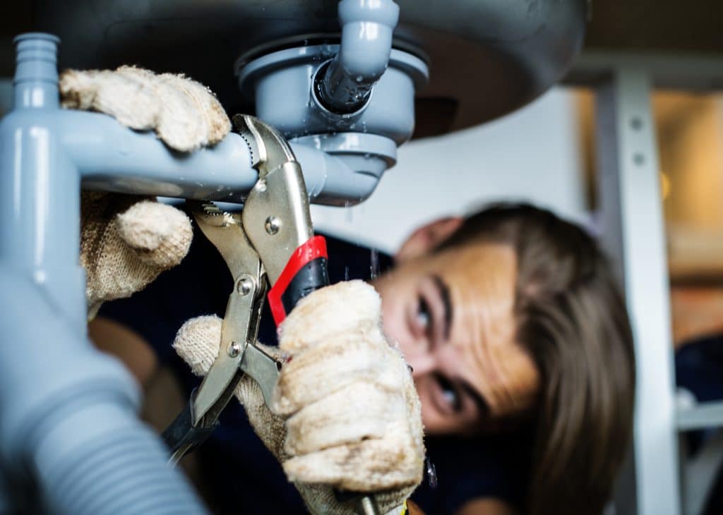 Gloved hands use locking pliers to tighten a leaking pipe joint under a sink, with water dripping from the plumbing