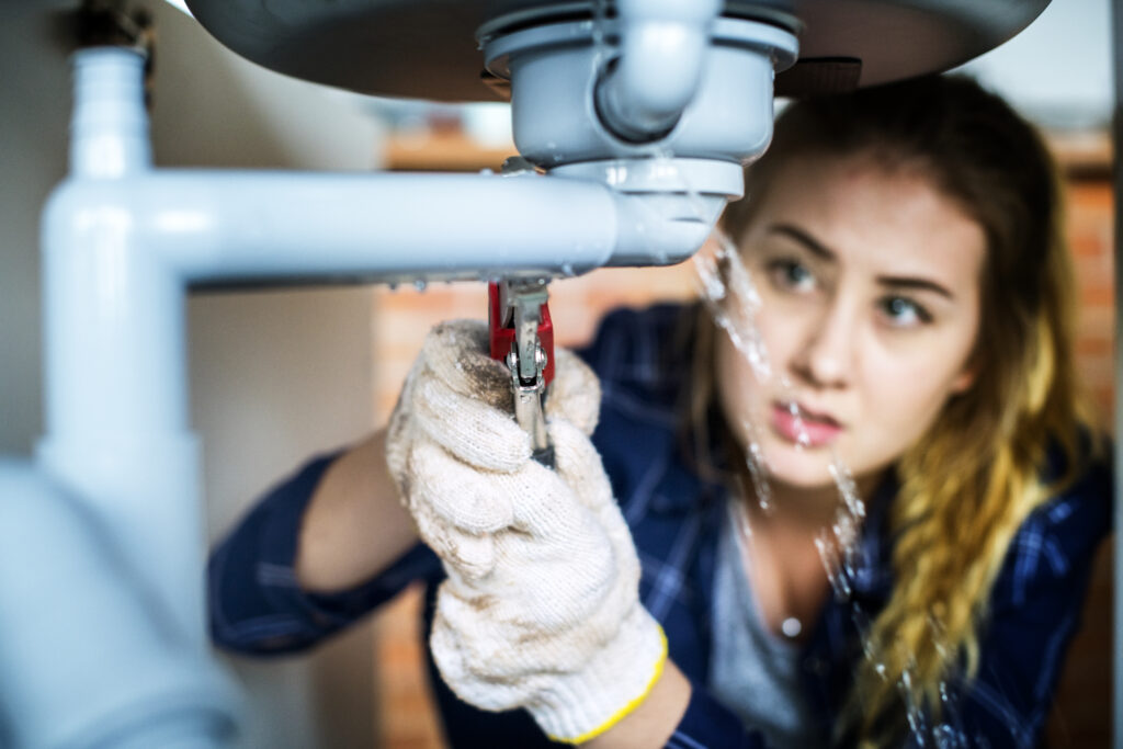 Woman tightening a leaking kitchen sink pipe with a wrench as water sprays under the sink