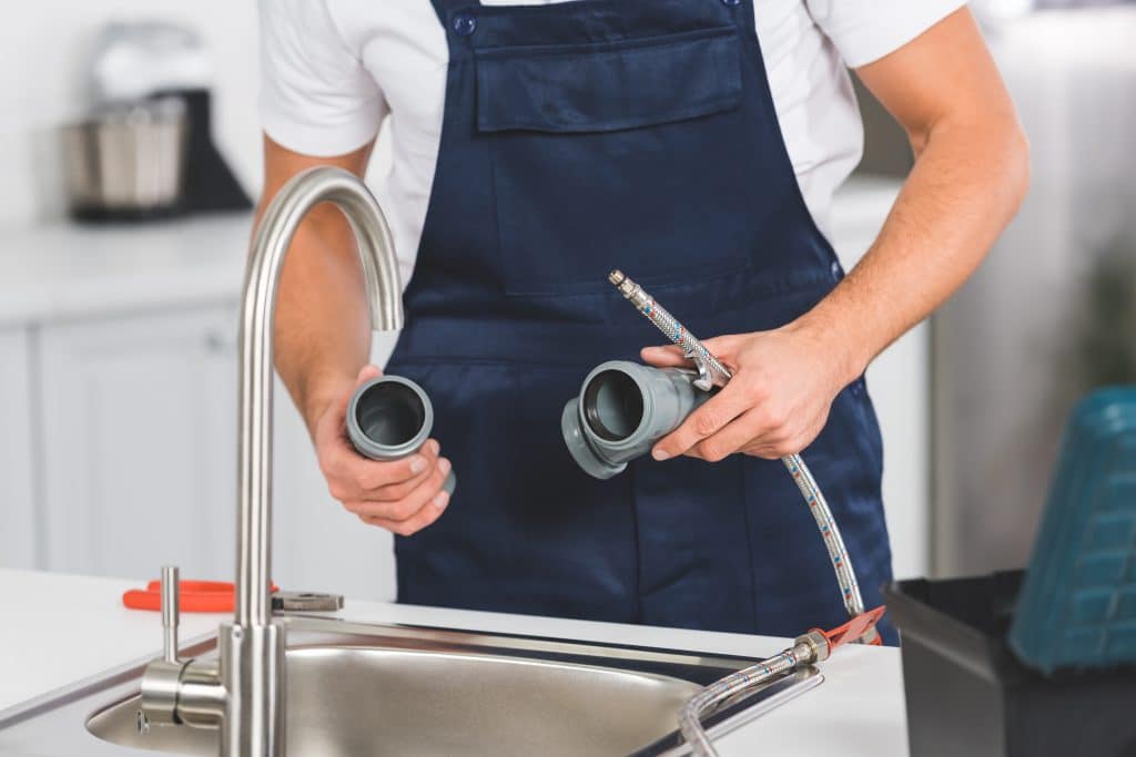 Cropped plumber holding PVC fittings and braided hose while repairing a kitchen sink faucet