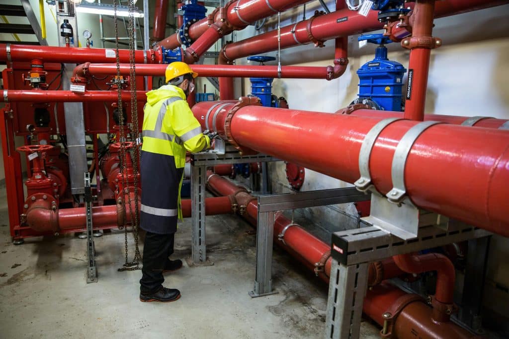 Worker in hard hat and high-vis jacket inspecting large red industrial pipes and blue valves in a mechanical room