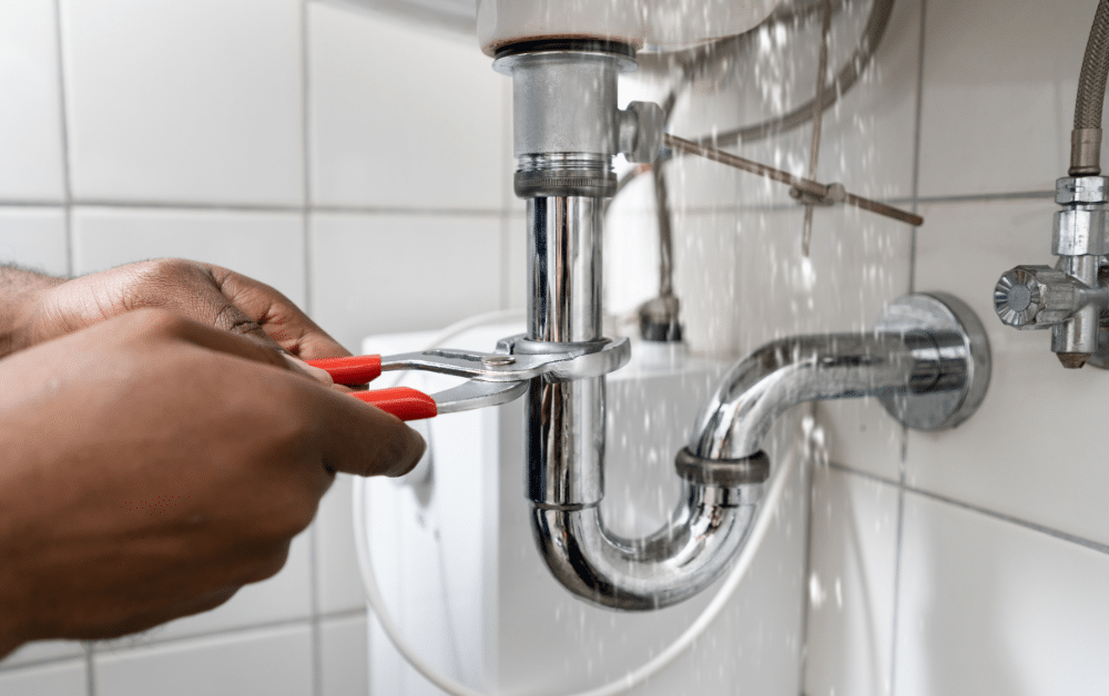 Hands using pliers to tighten a leaking sink drainpipe under a bathroom sink, with water spraying from the connection