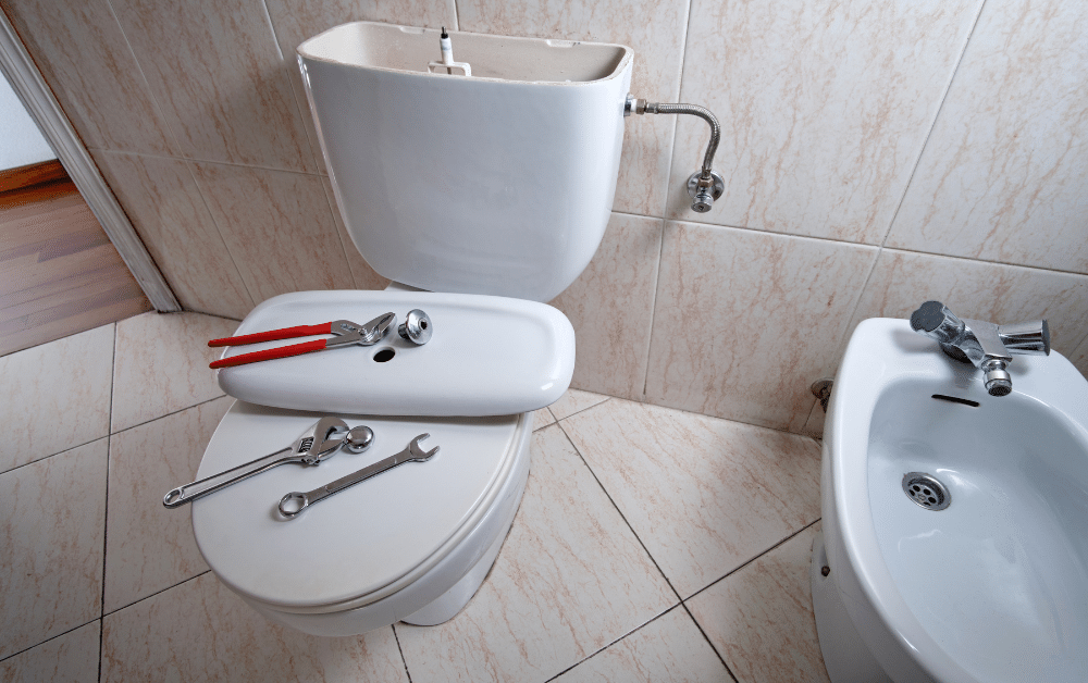 Toilet repair tools on tank lid and seat, with open cistern and nearby bidet in tiled bathroom