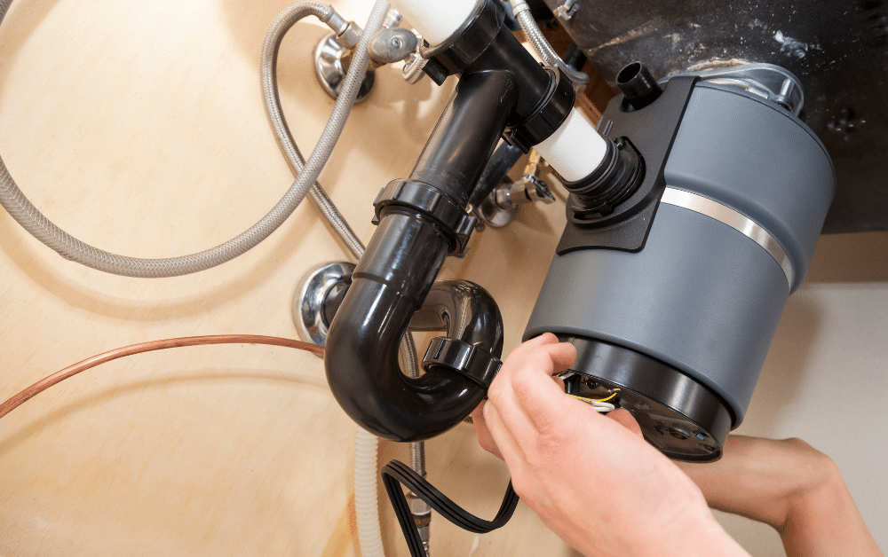 Hands installing a gray garbage disposal under a kitchen sink, connecting wiring and black drain pipe