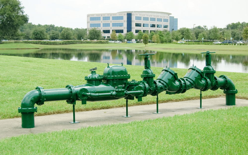 Green backflow preventer piping assembly on concrete pad beside a pond, with an office building in the background