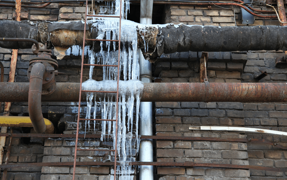 Frozen, leaking industrial pipe with icicles and ice buildup on a brick wall, with ladder and rusted plumbing lines